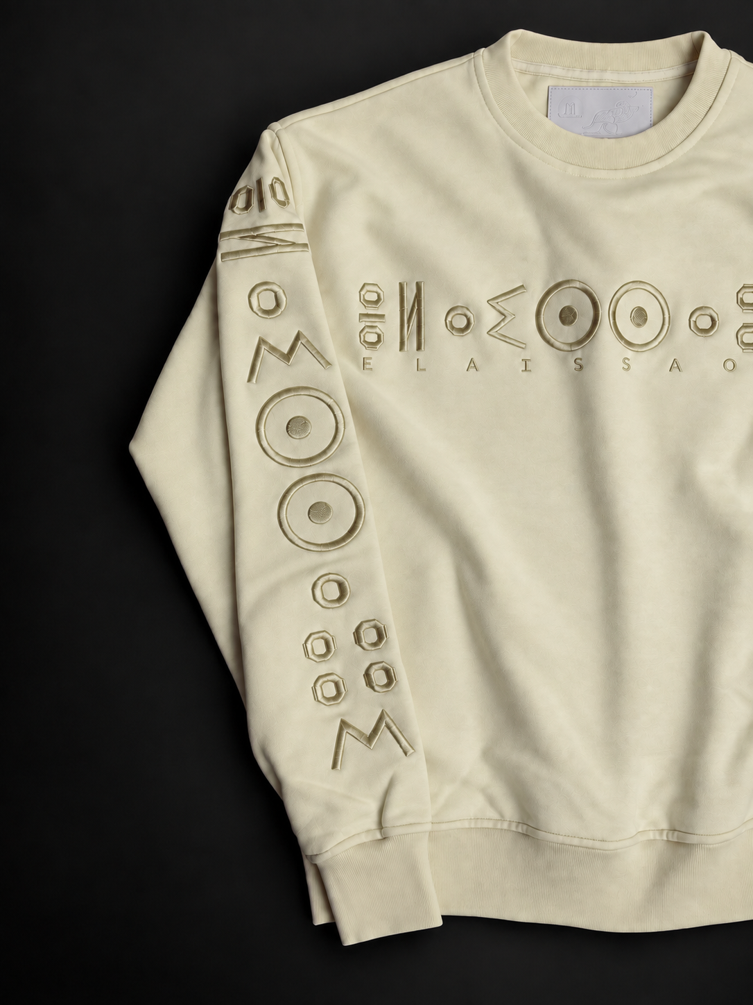 ELAISSAOUI TICHKA Desert Ivory pullover laid at an angle, showing the chest embroidery and the embroidered sleeve.